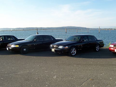 2003 Ford Crown Victoria - Several black sedans are parked along the waterfront on a sunny day.