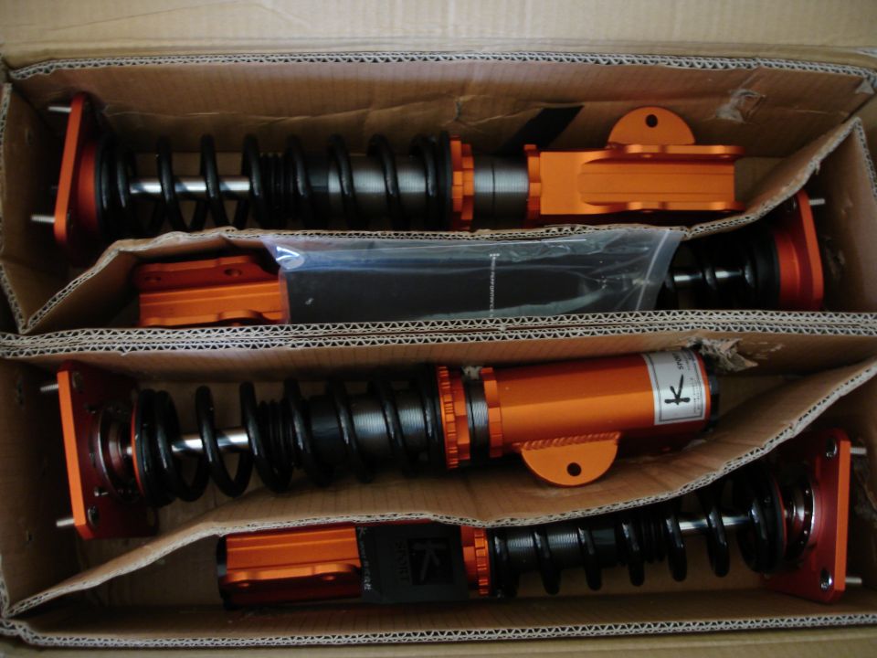 Four orange and black suspension coilovers in a cardboard box. Some have plastic bags