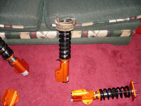 1989 Mazda MX-6 - Several orange suspension struts are laid out on a red carpet, with a couch visible behind them.