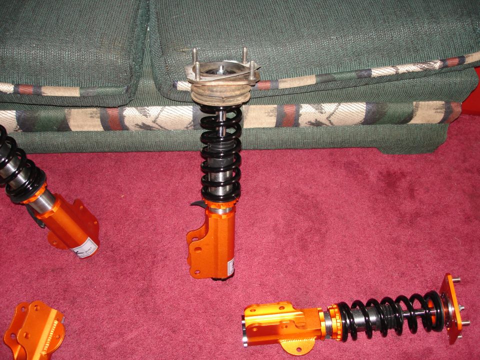 Three orange suspension struts with black springs on a red carpet, with a couch in the background