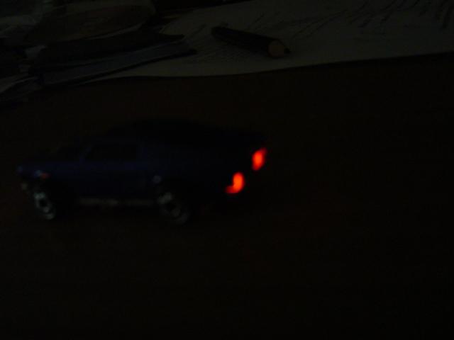 Dark blue toy car, rear view, with glowing red taillights. Supermotors.net watermark visible
