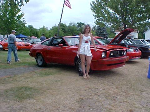 1978 Ford Mustang II - A woman poses next to a red 1978 Ford Mustang II convertible with its hood up at an outdoor car show.