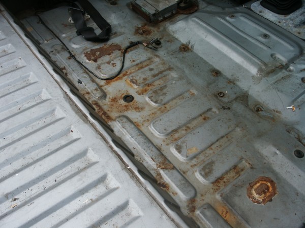 Rusted floor pan and seat mounting points inside a vehicle. Some wiring is visible