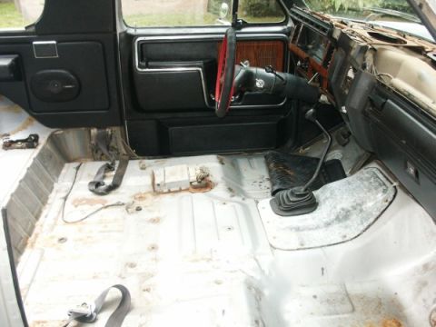 1982 Ford Bronco - The interior of a truck shows exposed metal flooring and a black door panel, with a red steering wheel visible.