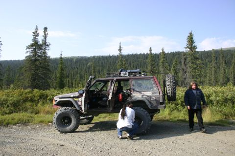 2003 Jeep TJ - A maroon Jeep Cherokee XJ is parked off-road with two people attending to it, surrounded by a forest.