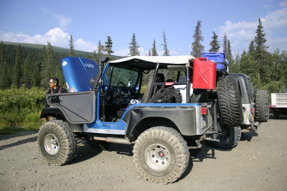 Blue Jeep CJ with hood open, spare tire mounted on rear, and jerry cans on rack. Off-road tires and suspension visible