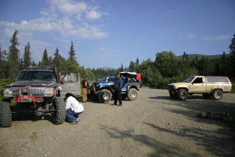 2003 Jeep TJ - A group of off-road vehicles are parked on a gravel lot with trees and mountains in the background.