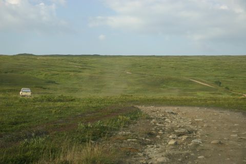 2003 Jeep TJ - A lone white SUV travels across a vast, green, rolling landscape under a partly cloudy sky.