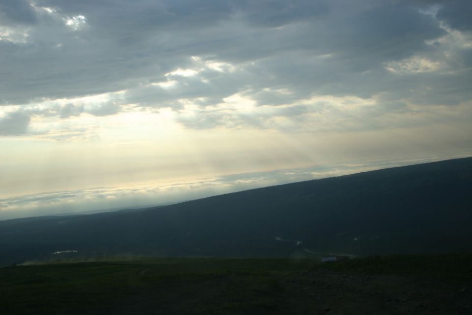 Overcast sky with sunbeams breaking through above a dark, tree-covered hillside