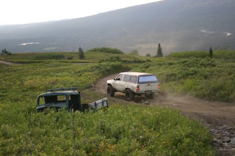 2003 Jeep TJ - A white SUV travels on a dirt road through a scenic landscape with an abandoned truck nearby.
