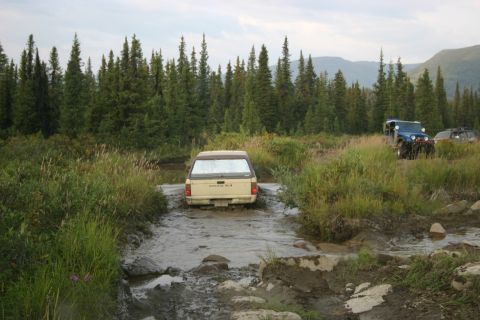 2003 Jeep TJ - Multiple off-road vehicles navigate a wet, wooded trail with a beige pickup truck in the foreground.