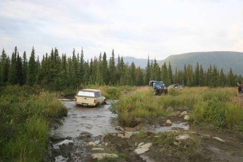 2003 Jeep TJ - Several off-road vehicles traverse a shallow, muddy stream in a wooded, mountainous area.