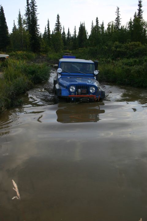 2003 Jeep TJ - A blue 2003 Jeep TJ navigates a muddy trail surrounded by dense forest.