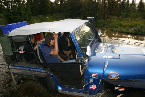 2003 Jeep TJ - A blue Jeep Wrangler navigates through a muddy, watery terrain, equipped with a snorkel and a roof rack carrying supplies.