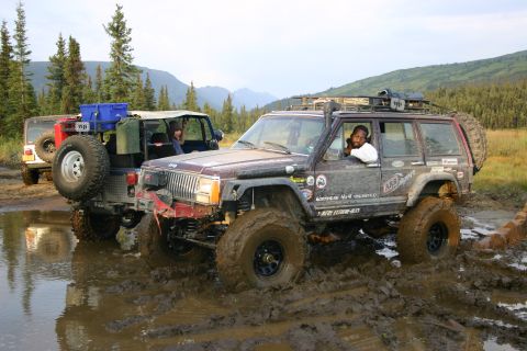 2003 Jeep TJ - Two modified Jeeps navigate a muddy off-road trail with mountainous terrain in the background.