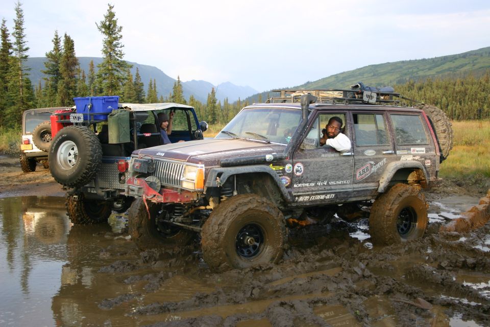 Mud-covered maroon Jeep Cherokee XJ with roof rack and snorkel stuck in mud, driver visible