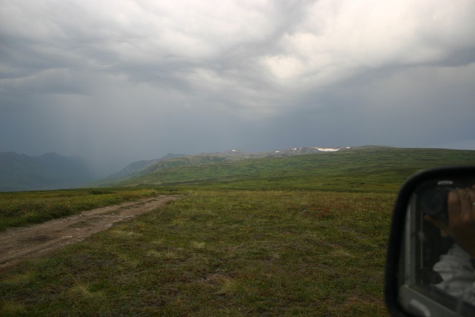 dirt road leading through grassy hills toward mountains under a stormy sky. Side mirror visible on right
