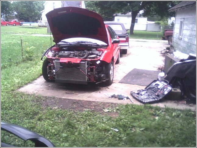 Red car with hood open, front bumper removed, parked outside