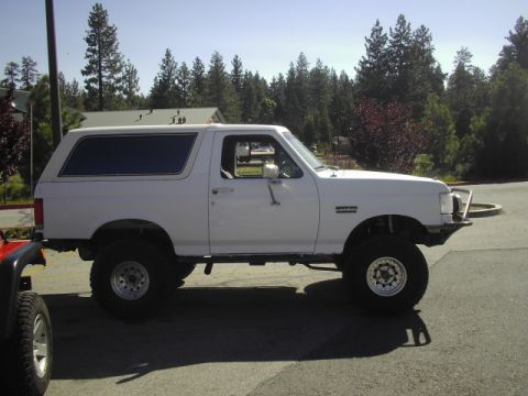 1990 Ford Bronco - white lifted Ford Bronco SUV with a tubular front bumper parked in a paved driveway by pine trees.