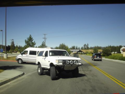 1990 Ford Bronco - white lifted Ford pickup with a winch bumper and round driving lights connected to a white van on a sunny road.