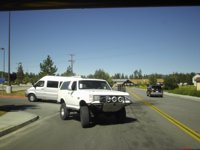 White lifted Ford pickup with auxiliary lights towing a white van on a road