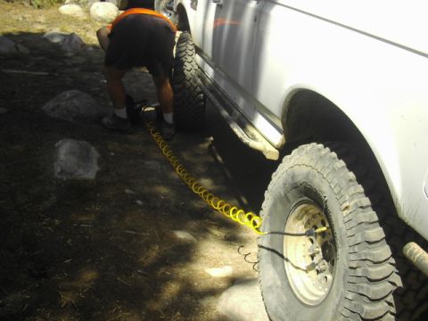 1990 Ford Bronco - person in an orange-strapped shirt crouches with a yellow air hose to deflate a tire on a muddy white off-road truck.