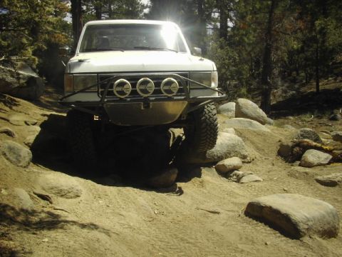 1990 Ford Bronco - white Ford off-road truck with three round driving lights crawling over large boulders on a rocky dirt trail.