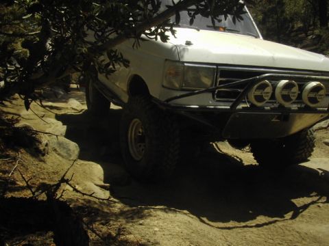 1990 Ford Bronco - white lifted Ford truck with a light bar climbs over rocks beneath a tree, one wheel articulated up on the trail.