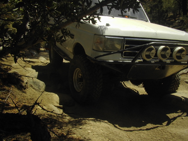 White lifted Ford truck flexing over rocks under a tree on an off-road trail