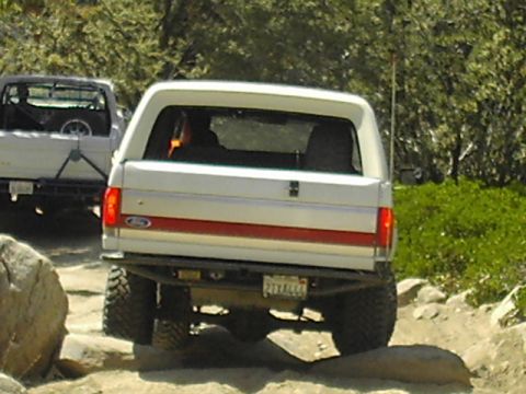 1990 Ford Bronco - Rear of a white Ford Bronco with a red stripe and Ford tailgate badge crawling over rocks, another truck behind it.