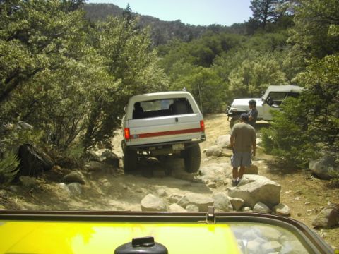 1990 Ford Bronco - white Ford SUV and other off-road trucks stopped on a rocky forest trail, with people standing among the rocks.