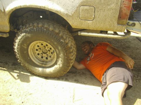 1990 Ford Bronco - smiling man in an orange shirt lies on the ground next to the large knobby tire of a mud-covered off-road truck.