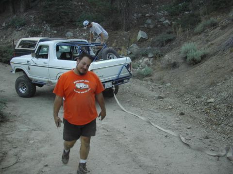 1990 Ford Bronco - man in an orange shirt walks toward the camera from a white Ford off-road truck with its winch line extended on a trail.