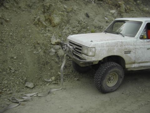 1990 Ford Bronco - heavily mud-splattered white Ford off-road truck nosed up against a rocky bank, a recovery strap lying on the dirt.