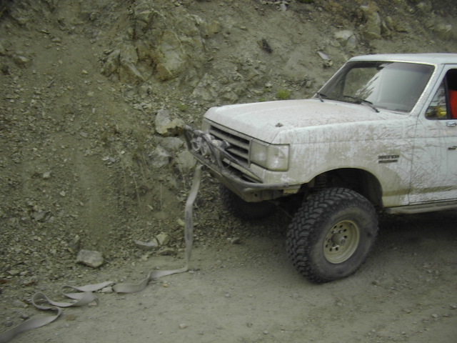 Mud-splattered white Ford truck nosed against a rock wall with a strap and winch
