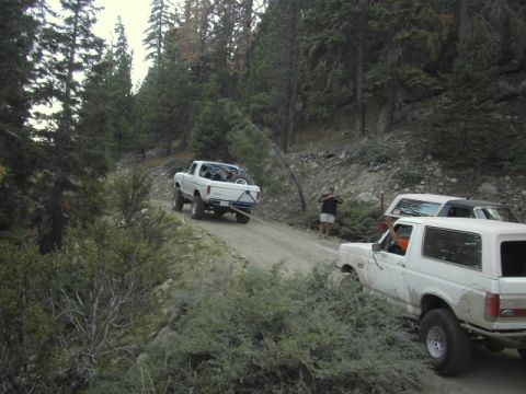 1990 Ford Bronco - white pickup climbs a narrow mountain dirt road while a person spots; a white Ford Bronco waits in the foreground.