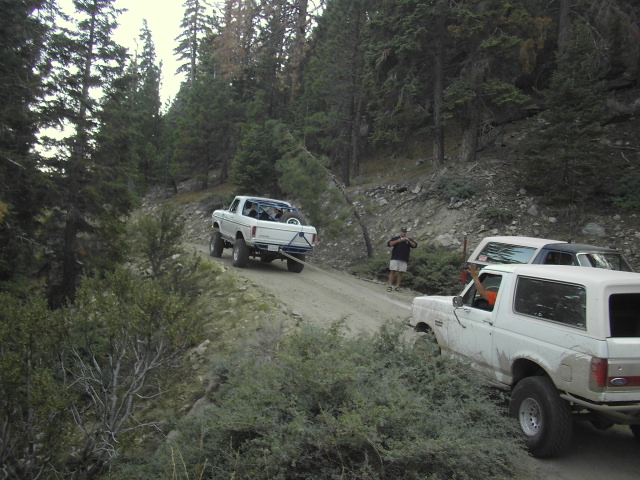 White pickup and white Bronco on a narrow mountain dirt road with a spotter