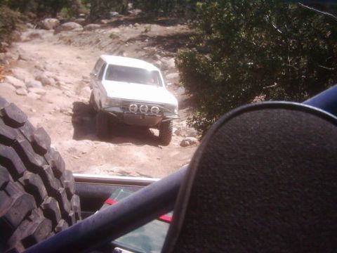 1990 Ford Bronco - white off-road truck with three round driving lights sits on rocky terrain, framed past a spare tire and seat back.