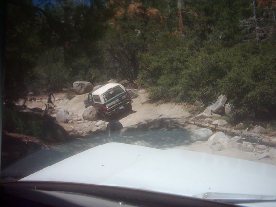 View through windshield of a white SUV descending a rocky off-road trail in pine forest