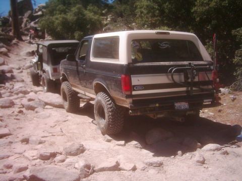 1990 Ford Bronco - lifted dark Ford Bronco with white roof and tire-carrier crawls up a boulder-strewn trail, another rig ahead of it.