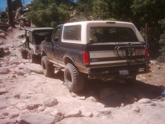 Rear of a lifted dark Ford Bronco with white top climbing a rocky trail