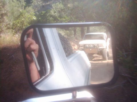 1990 Ford Bronco - side-mirror reflection captures a hand giving a thumbs-up while a lifted white truck with off-road lights follows on a dirt forest trail.
