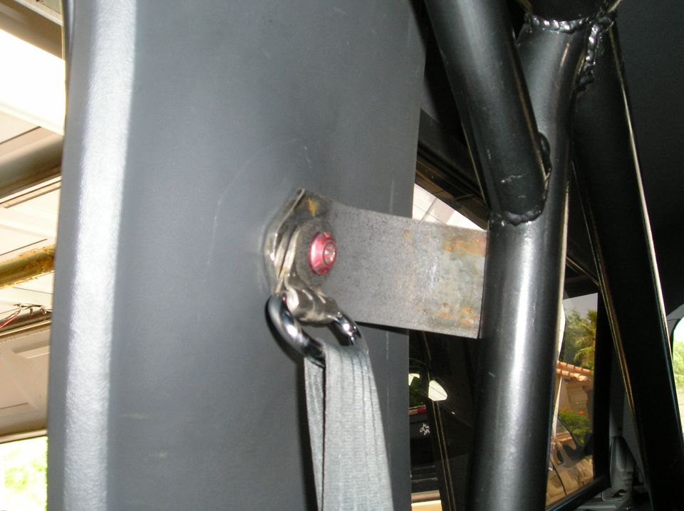 Seat belt mount with red bolt and metal bracket on gray pillar near black roll bar