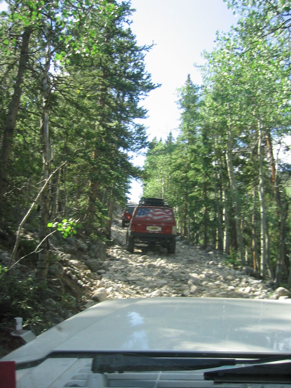 Red 1997 Jeep Cherokee with American flag decal on rear door, driving on a rocky mountain trail