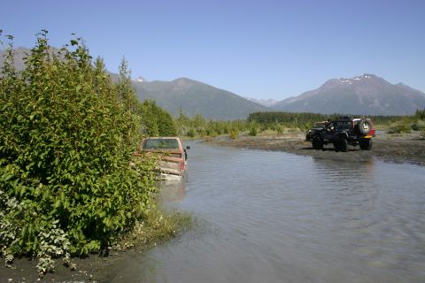 2003 Jeep TJ - Two off-road vehicles navigate a shallow riverbed with mountainous terrain in the distance.
