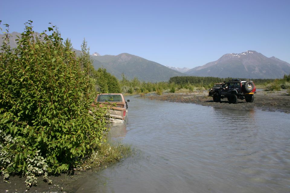 Red pickup truck partially submerged in water, with a dark SUV parked on the bank in the background. Mountains and trees form the backdrop