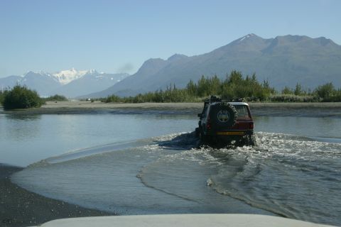 2003 Jeep TJ - A red SUV navigates a shallow river crossing under a clear blue sky with mountains in the distance.