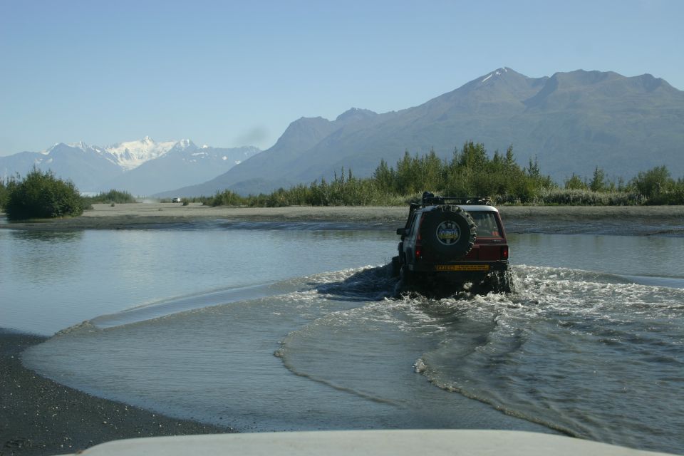 Red SUV crossing shallow river with snow-capped mountains in background