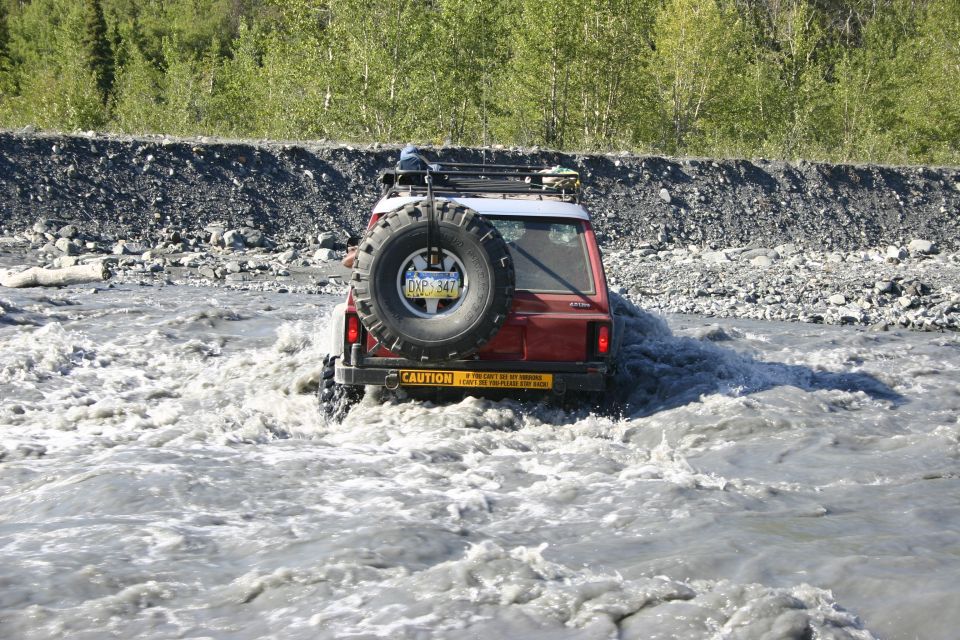 Red 2003 Jeep TJ driving through shallow water, spare tire mounted on rear door, roof rack visible