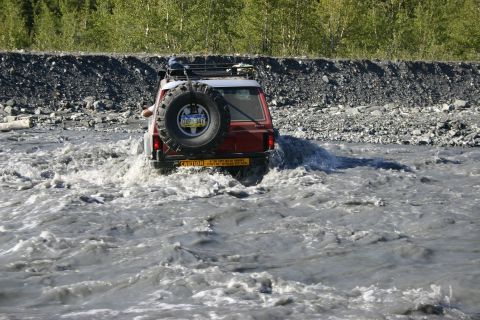 2003 Jeep TJ - A red SUV navigates a rocky riverbed, kicking up spray as it goes.